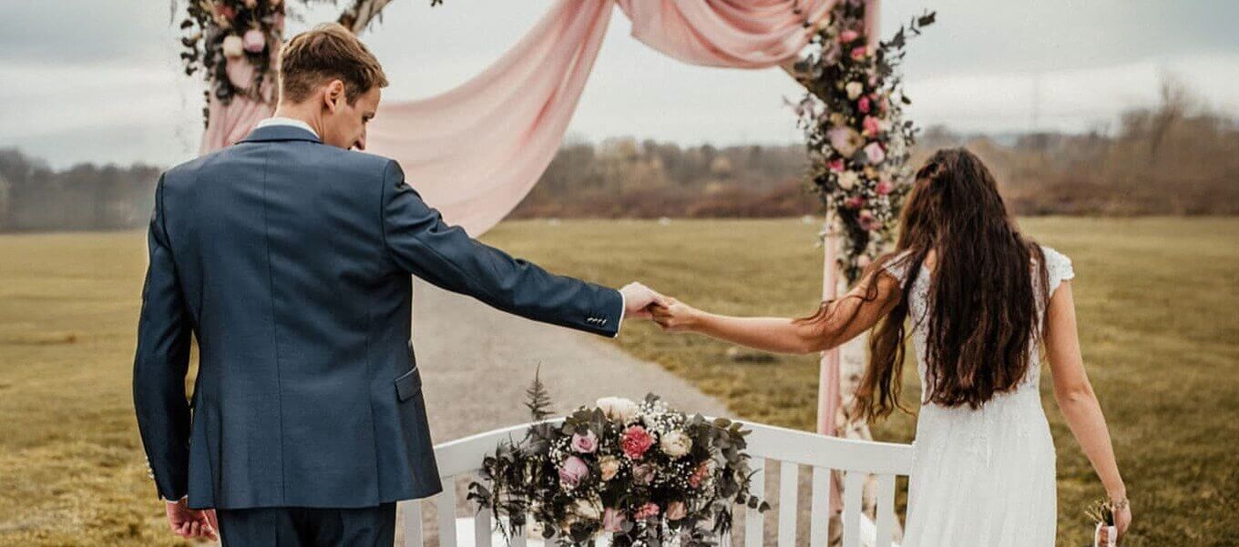Braut und Bräutigam laufen Hand in Hand durch eine Hochzeitslocation mit Blumen und dekoriertem Traubogen